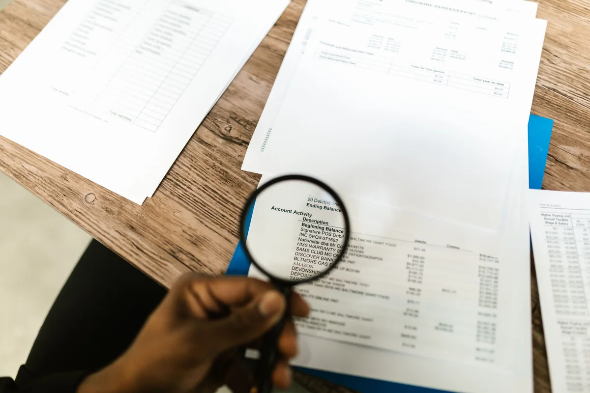 A hand holding a magnifying glass over a bank statement, with account activity and figures fully legible